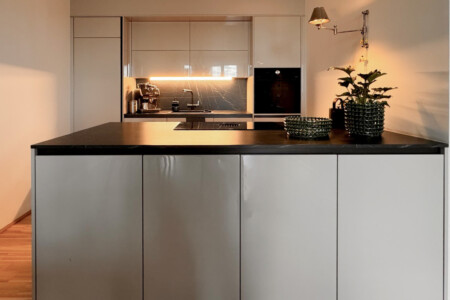 Contemporary kitchen with glossy beige island, black stone countertop, marble backsplash and a swing-arm wall lamp